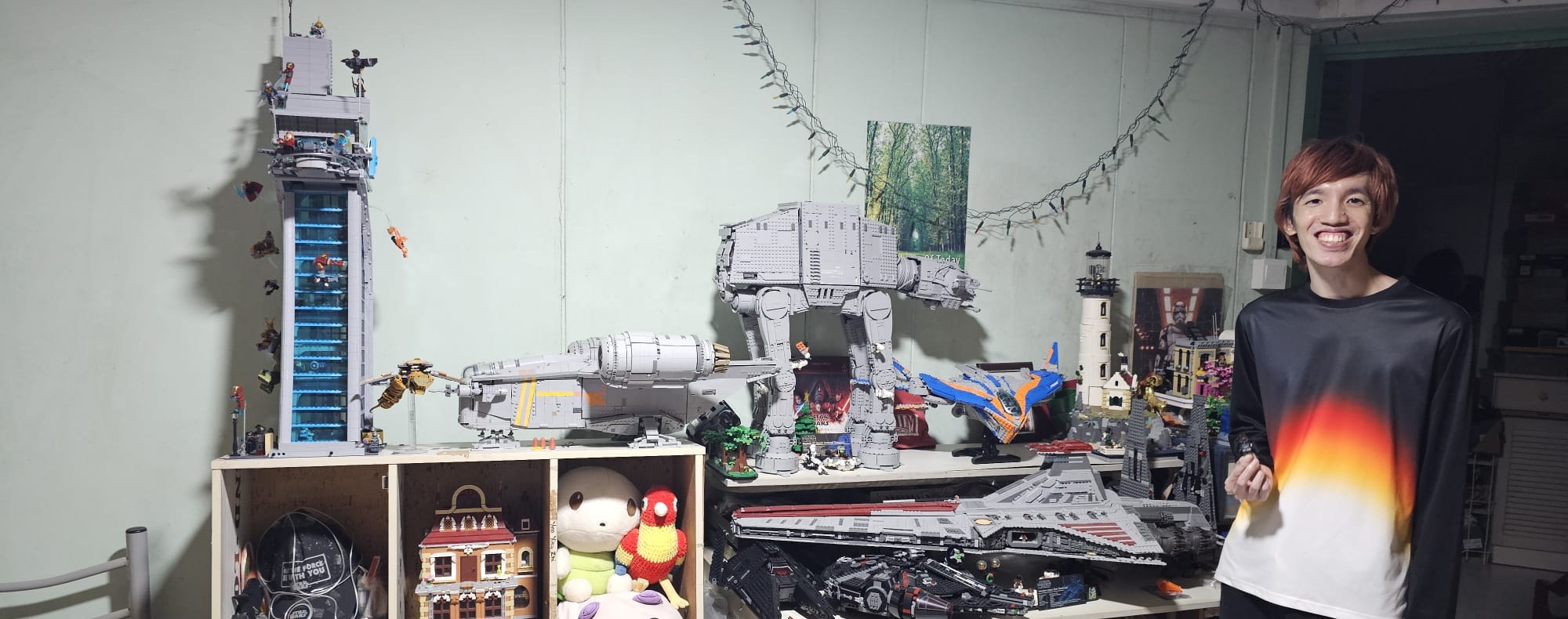 A founder standing beside a desk and shelves of brick builds: a tall tower, AT-AT walkers and a large grey starship, with string lights on the wall