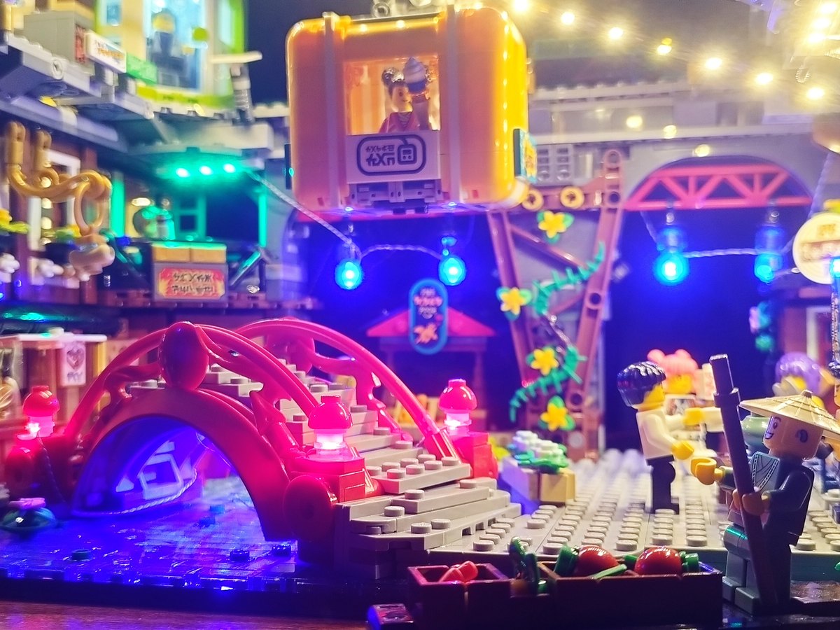 A red arched bridge and an orange gondola cabin among neon-lit market stalls, with string lights overhead